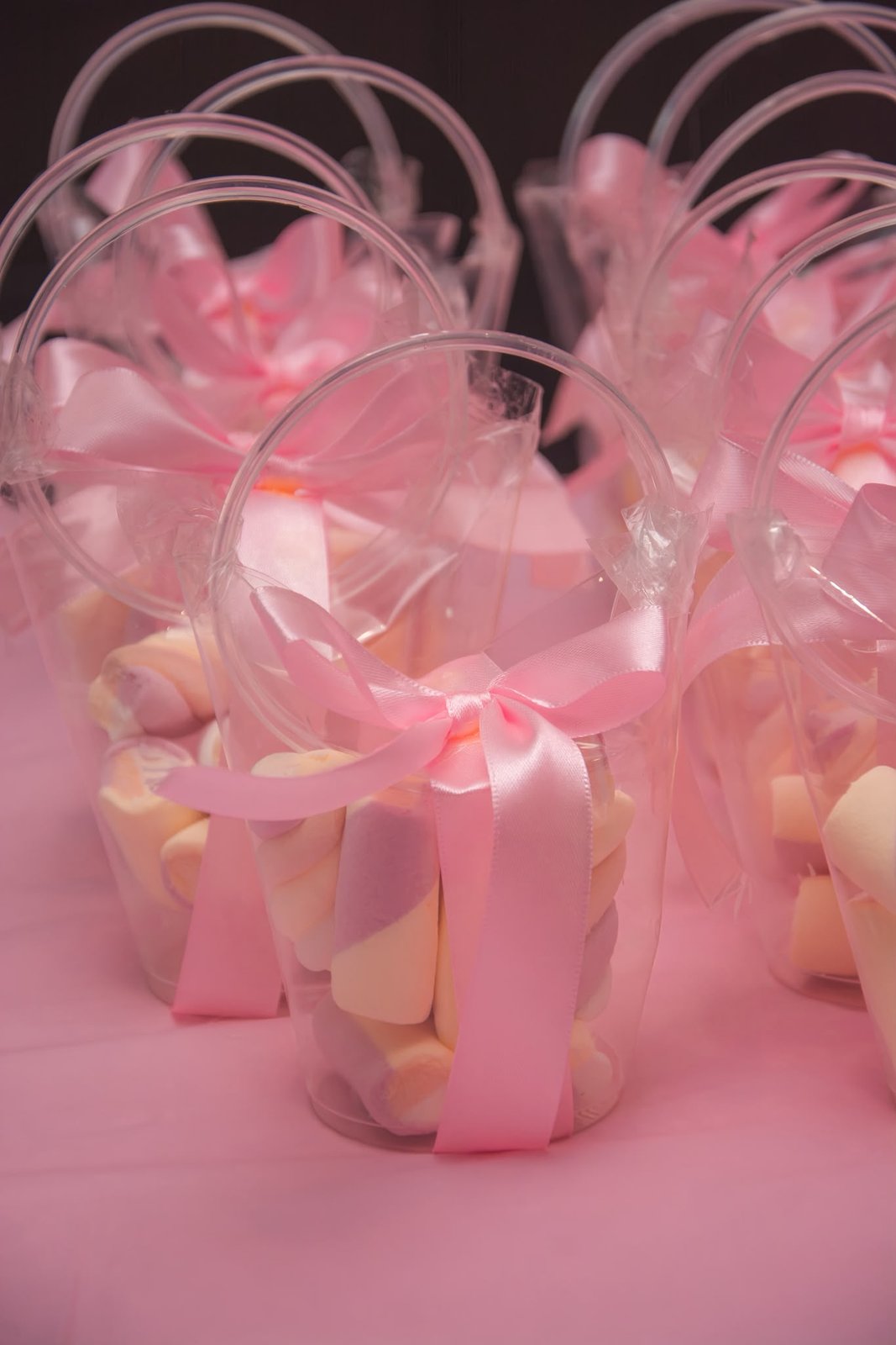 Cones transparentes com doces e laço de cetim rosa, alinhados sobre mesa rosa